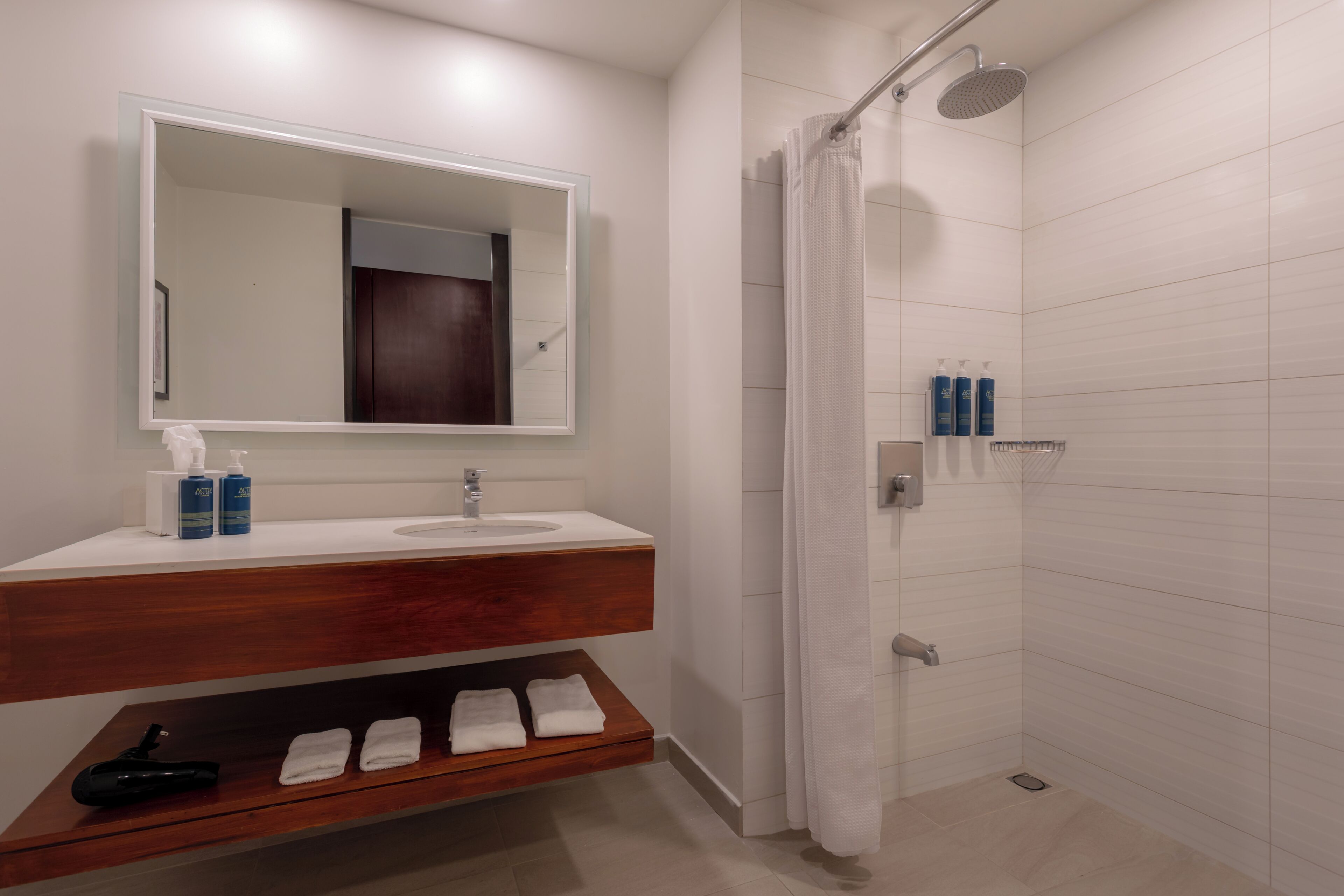 A bathroom with a wooden vanity, a large mirror, a shower with a curtain, and a shelf with toiletries.