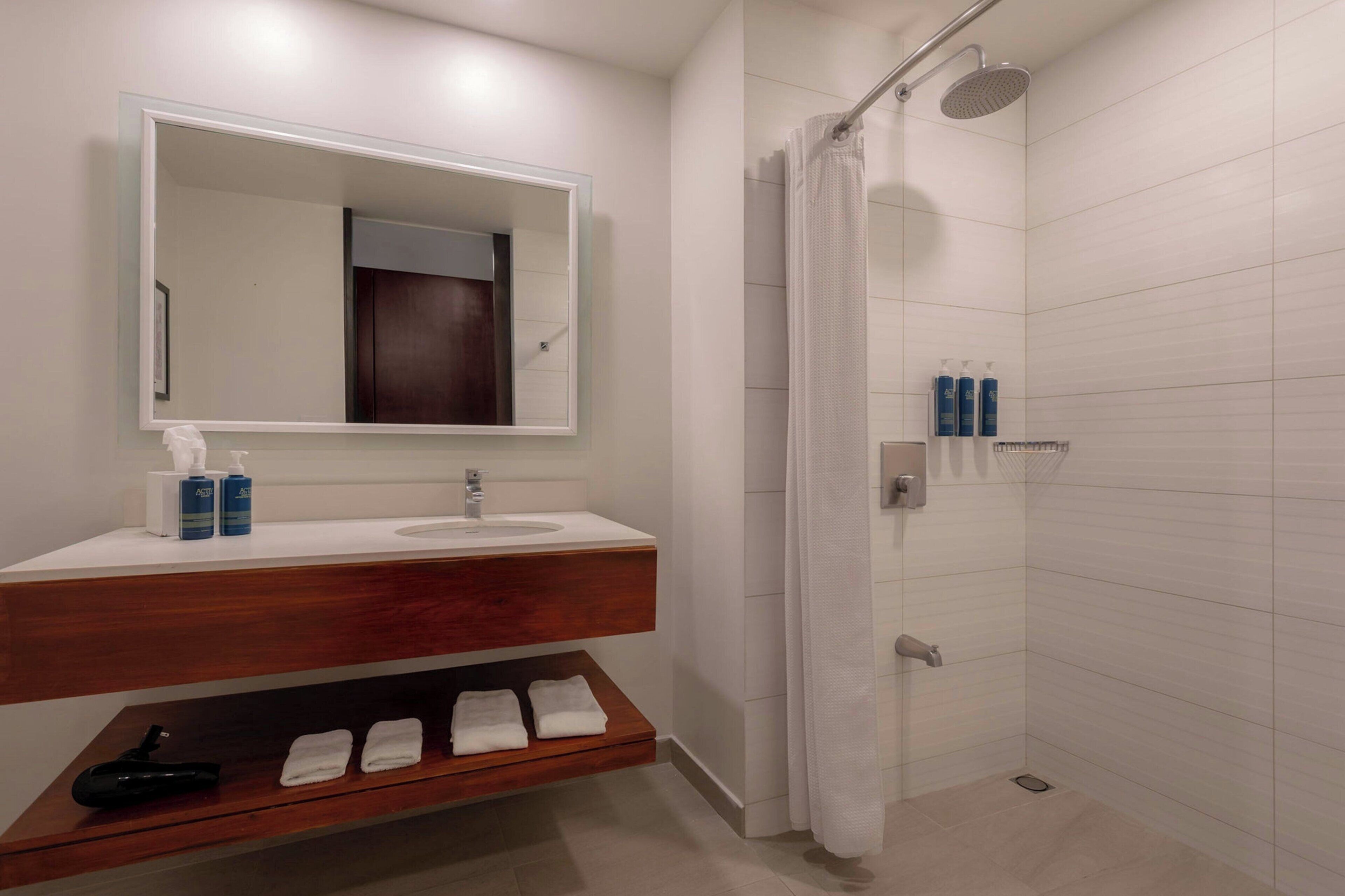 Un baño con lavamanos blanco, espejo y ducha. Hay toallas y artículos de aseo en la encimera.