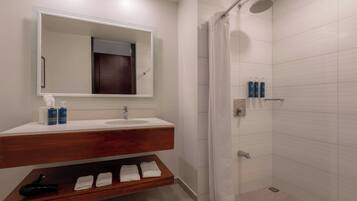 Un baño con lavamanos blanco, espejo y ducha. Hay toallas y artículos de aseo en la encimera.