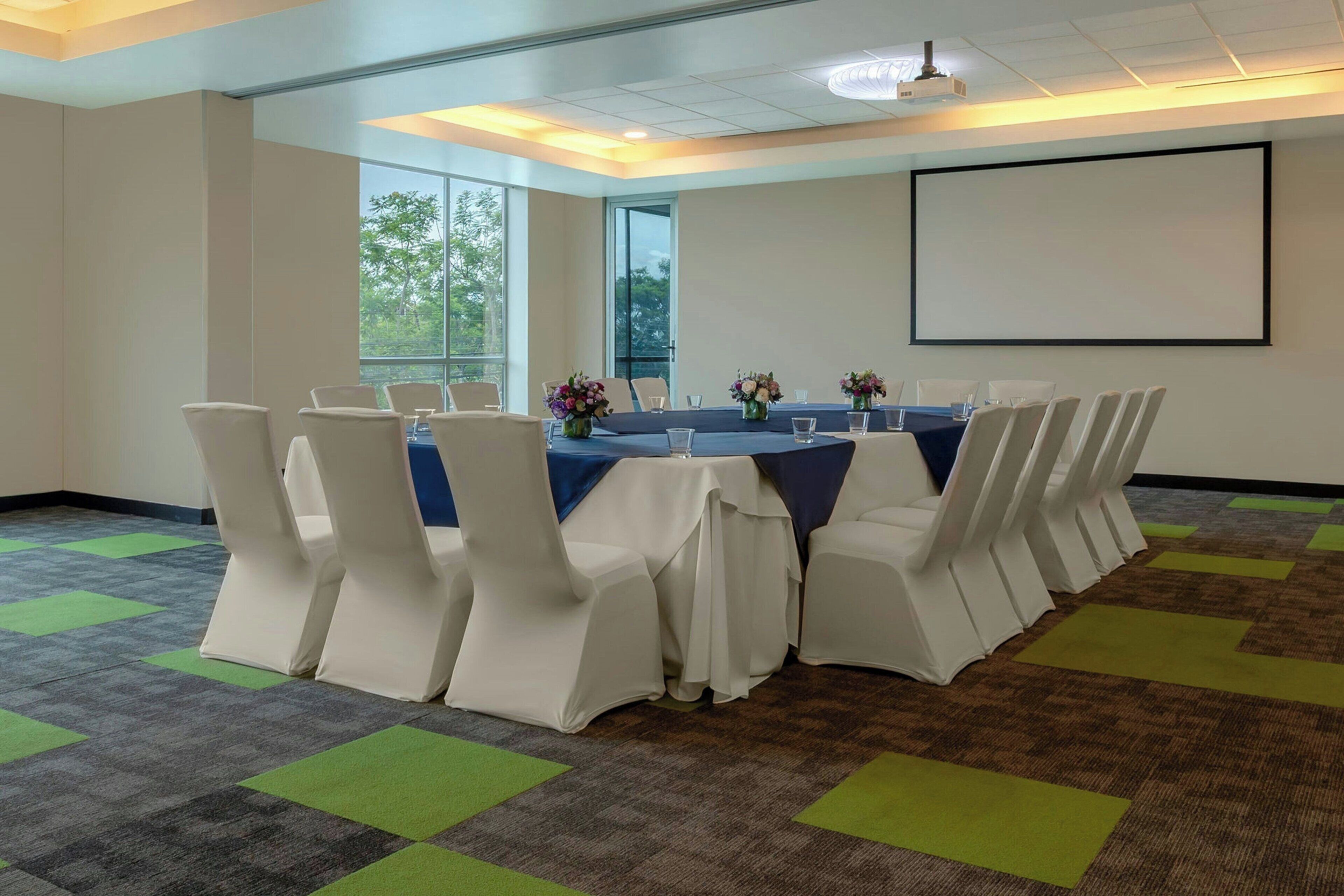 A conference room with a long table, white chairs, and a projector screen.