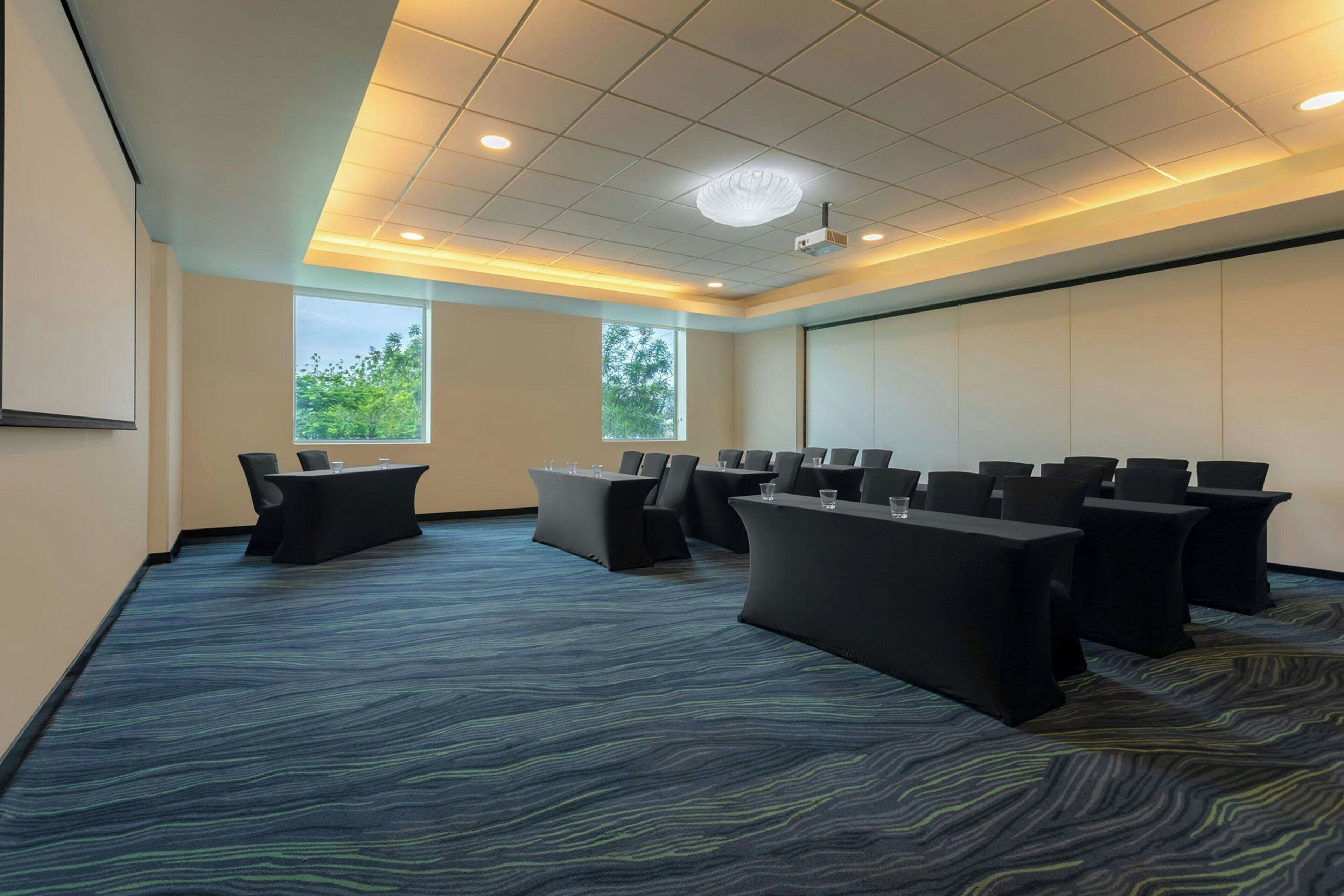 A conference room with a large screen, multiple tables with chairs, and two windows with a view of trees.
