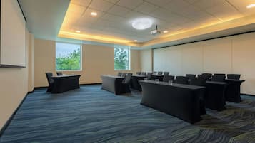 A conference room with a large screen, multiple tables with chairs, and two windows with a view of trees.