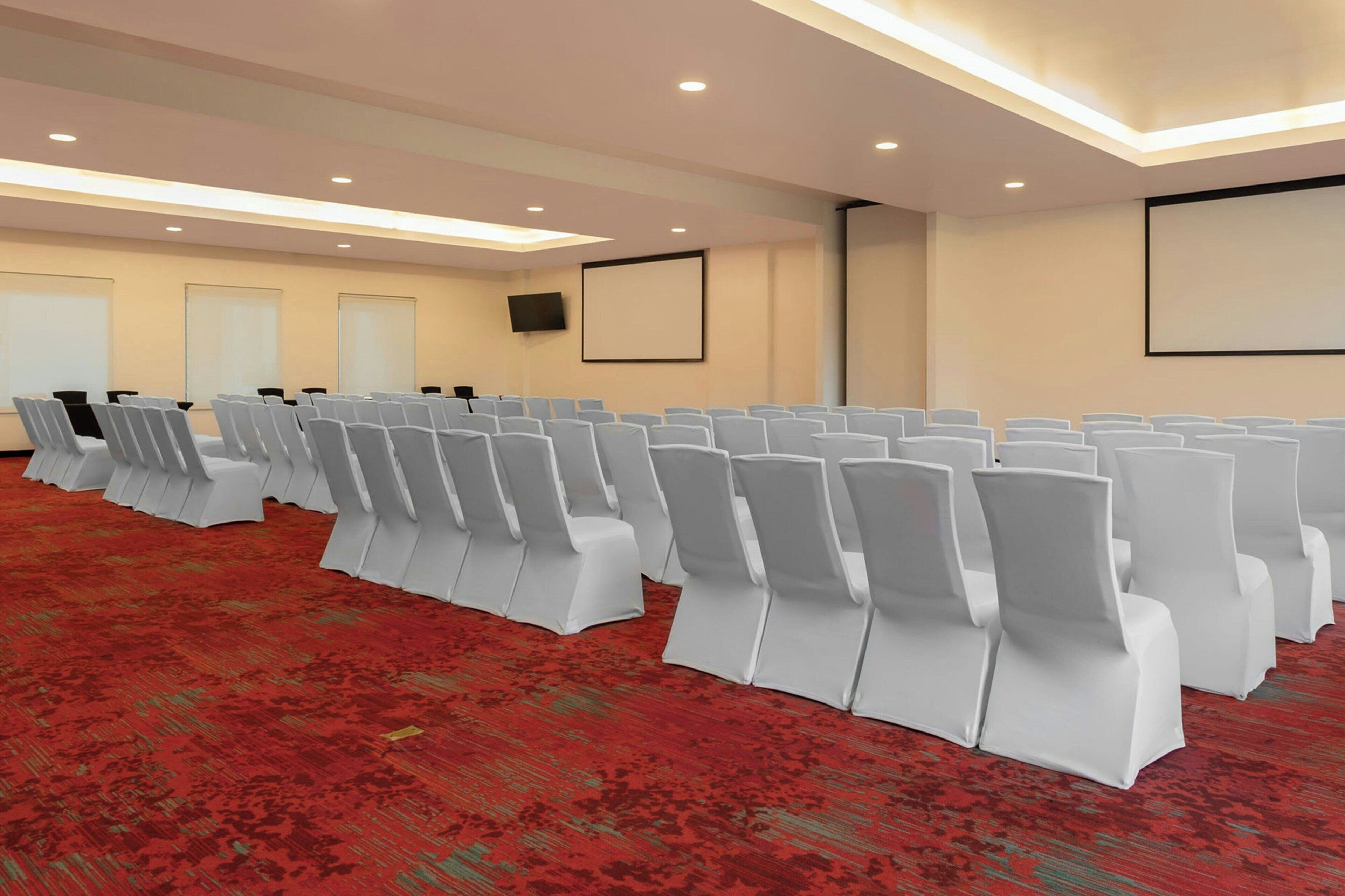 Un salón de conferencias con filas de sillas blancas, una alfombra roja, una pantalla de proyector y un televisor de pantalla plana en la pared.
