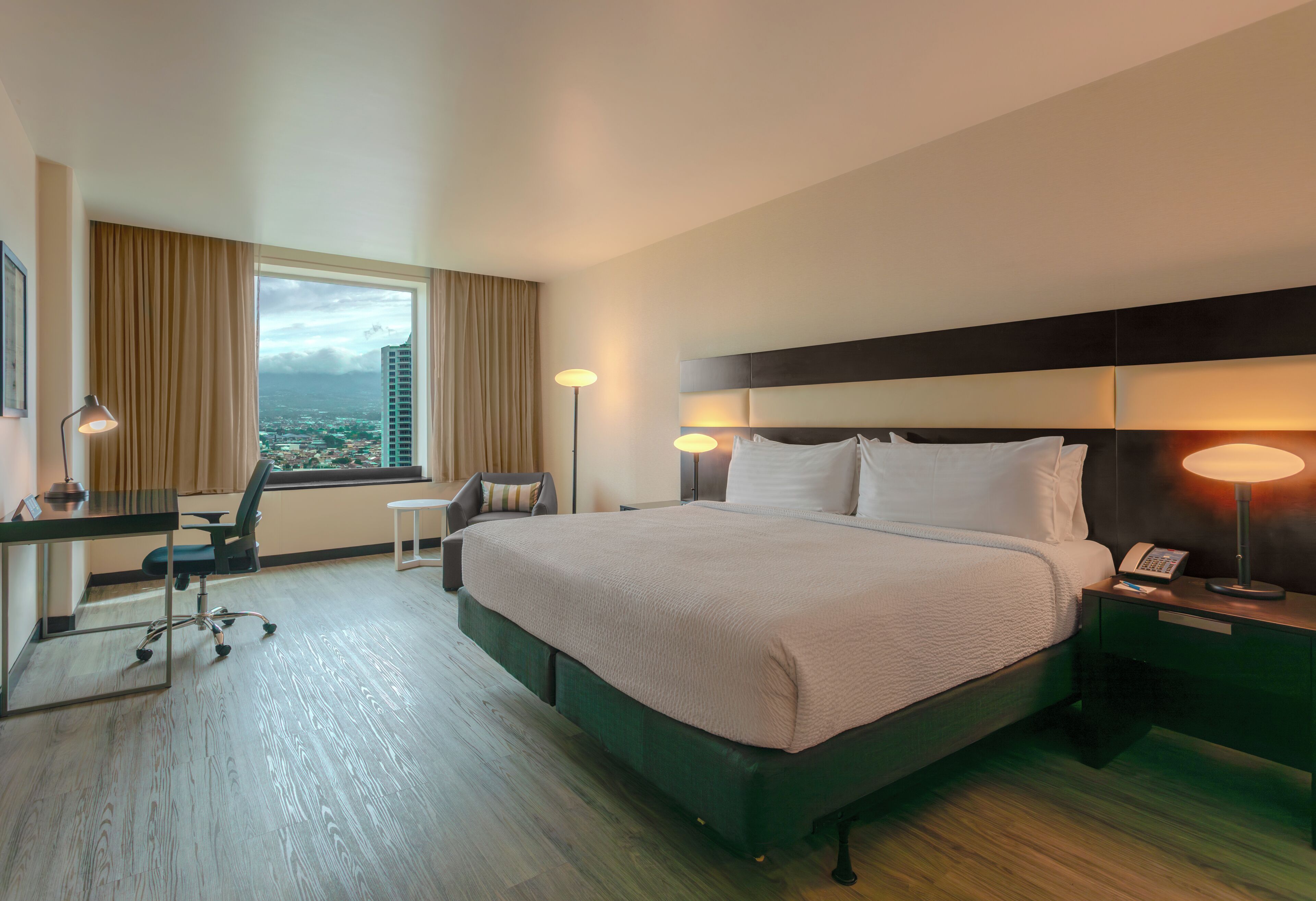 A hotel room with a bed, desk, chair, bedside table, lamp, and a view of a cityscape through the window.