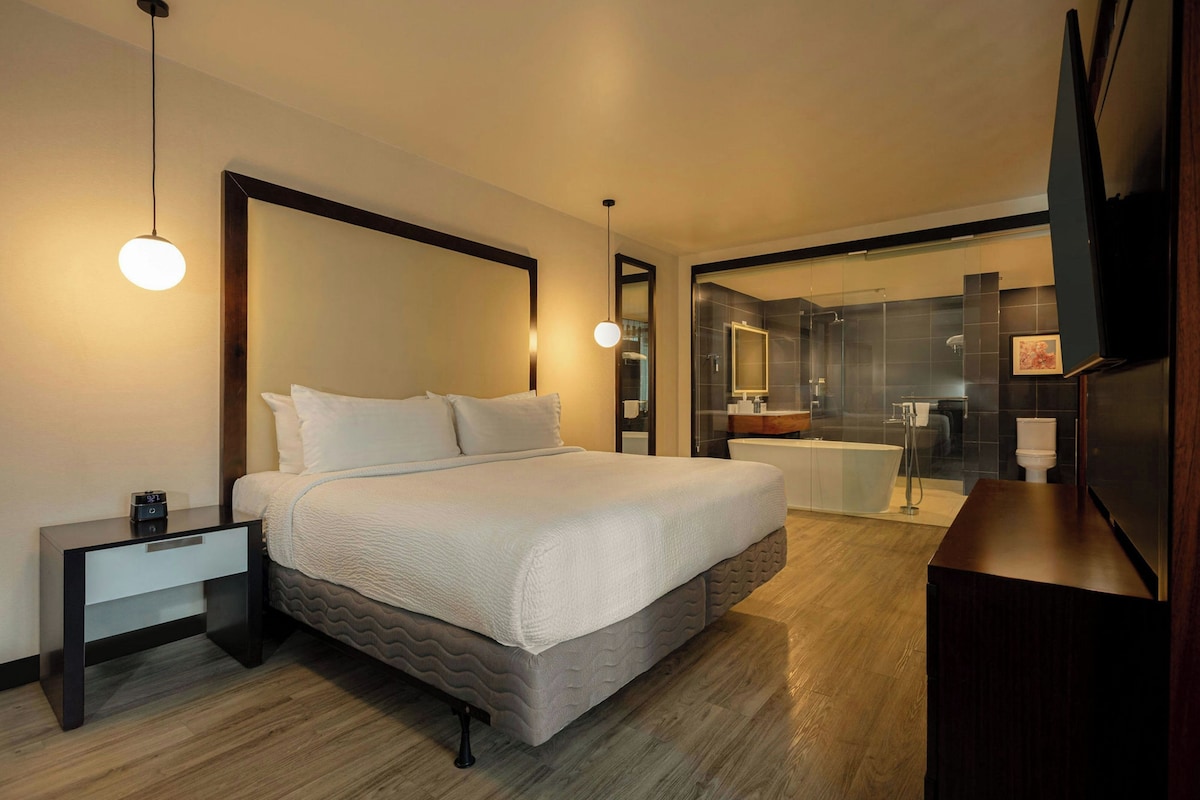 Une chambre d’hôtel moderne avec un grand lit, des tables de chevet, un plancher en bois, une salle de bain avec baignoire et un grand miroir.