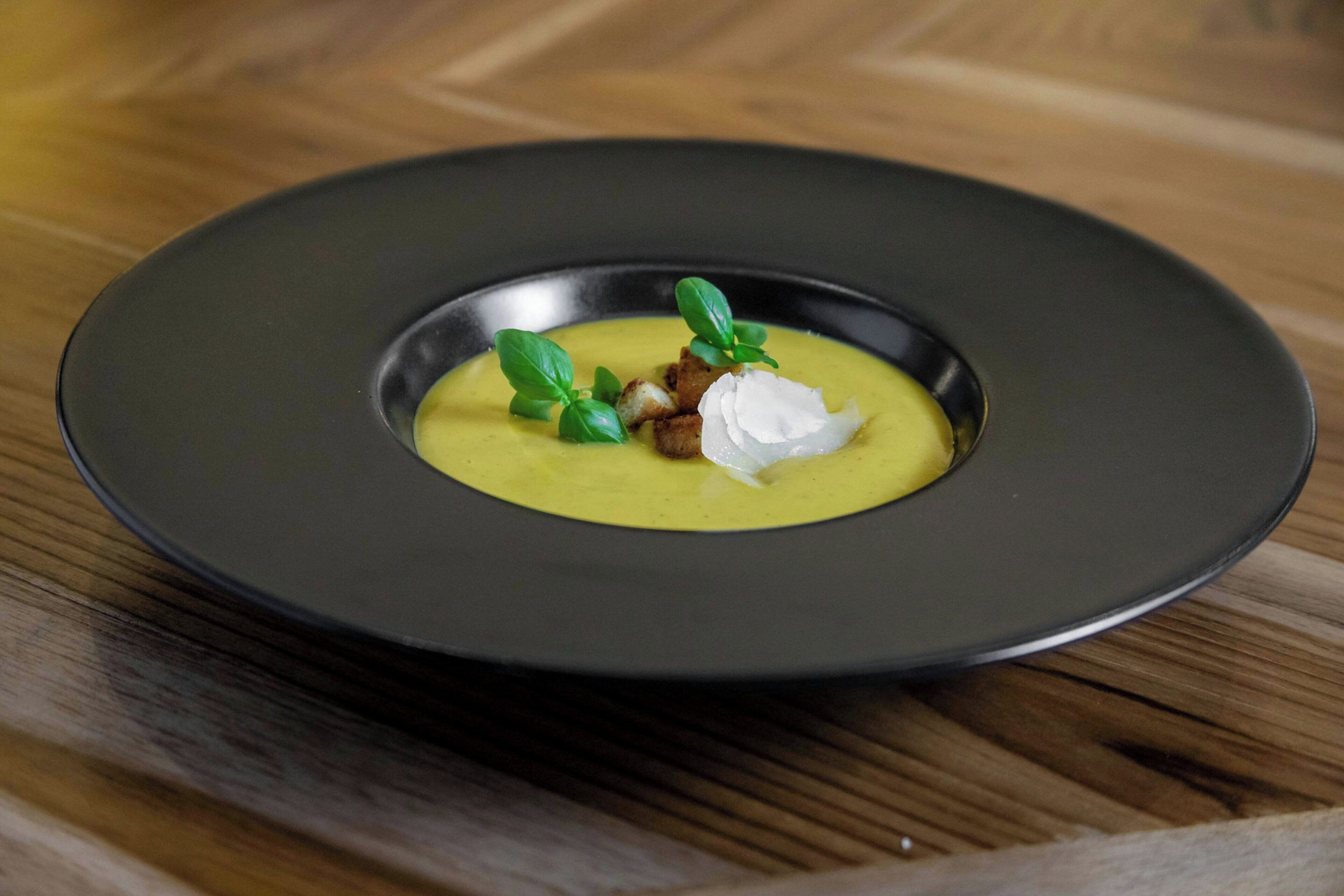 Een bord eten met een gladde, crèmige gele soep, versierd met een toef room en een takje groene kruid, op een donker bord.