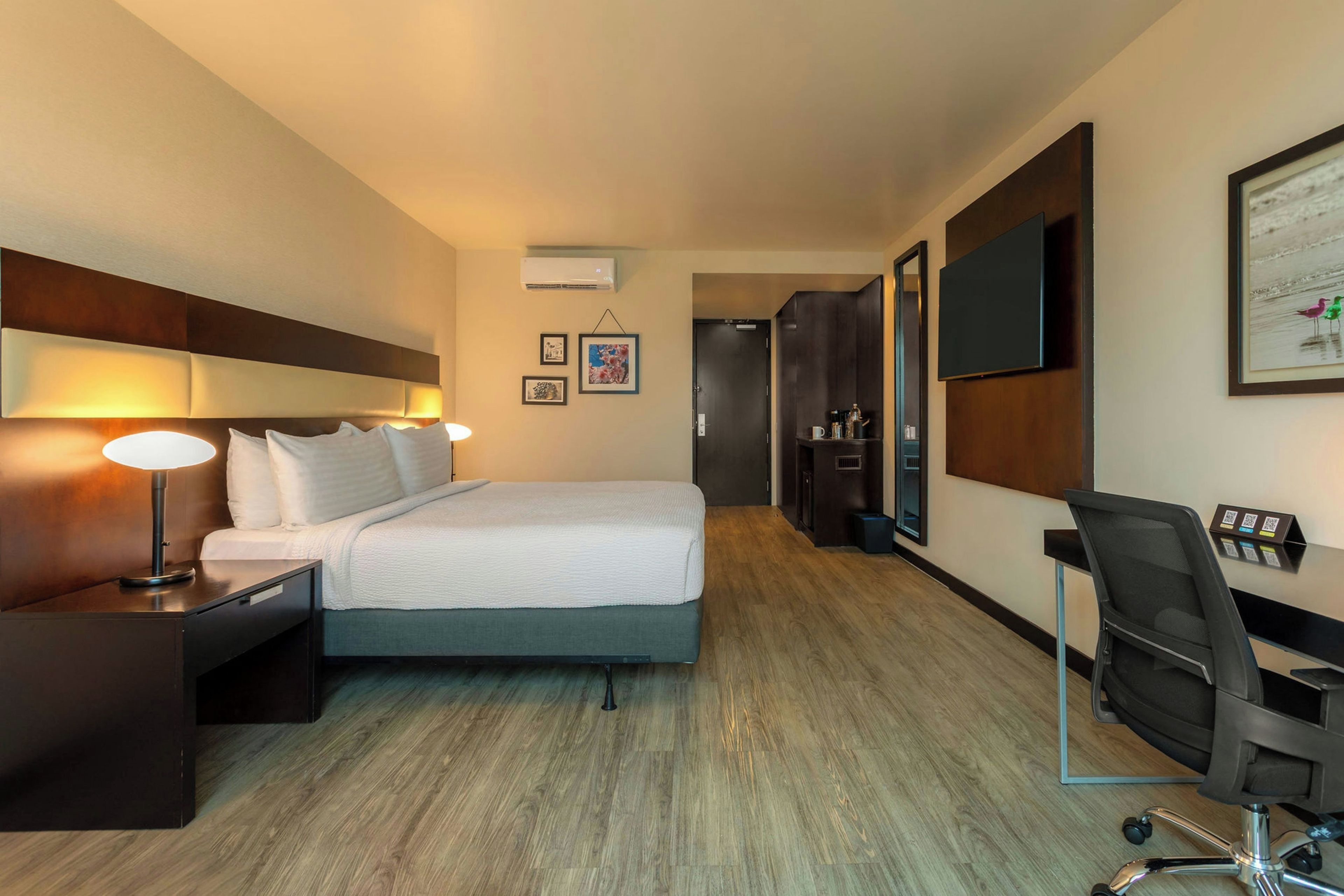 A hotel room with a bed, bedside tables, a desk with a chair, a flat-screen TV, and a framed picture on the wall.