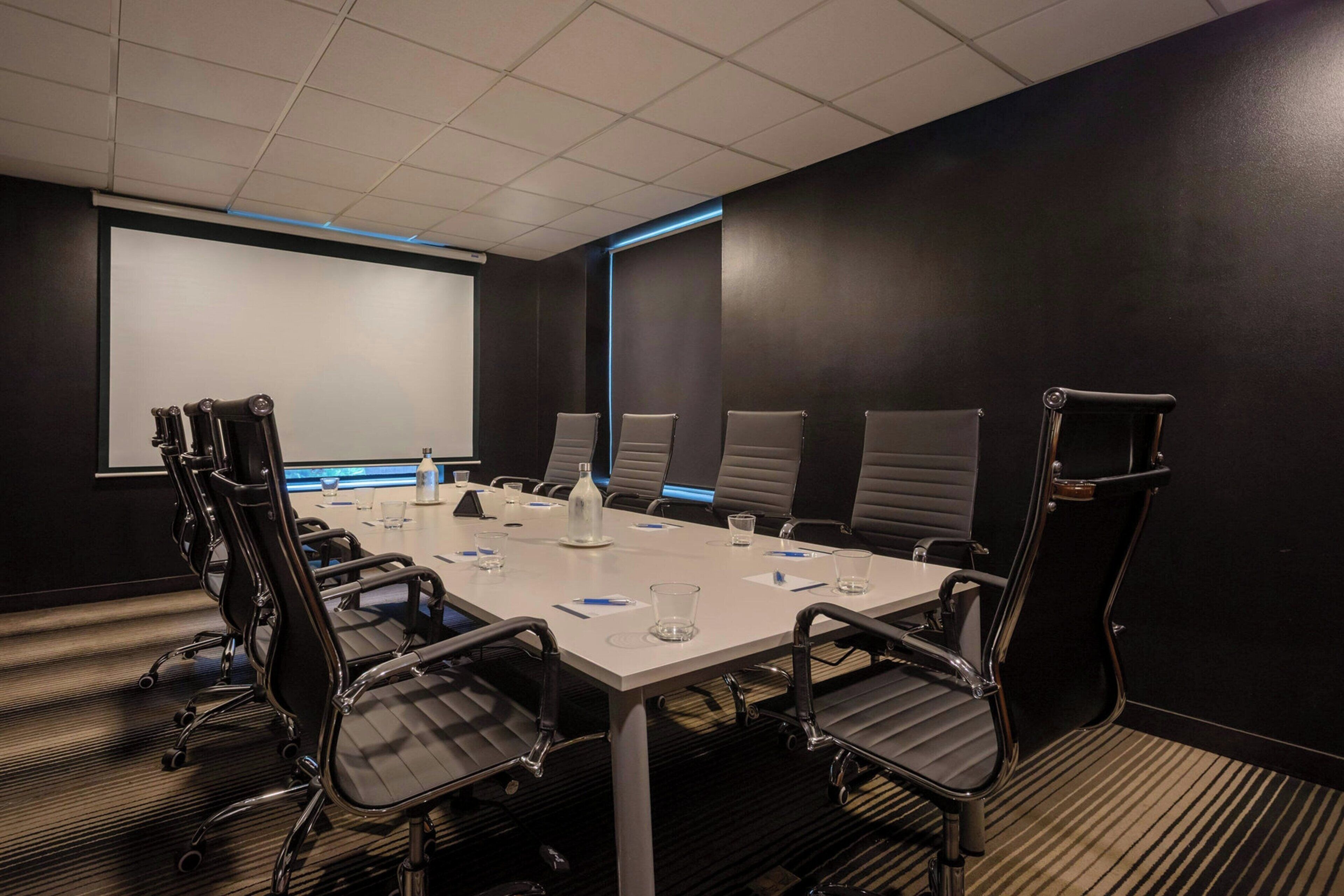 A conference room with a white table, black chairs, a projector screen, and a carpeted floor.