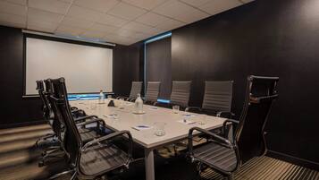 Un salón de conferencias con una mesa blanca, sillas negras, una pantalla de proyector y piso alfombrado.