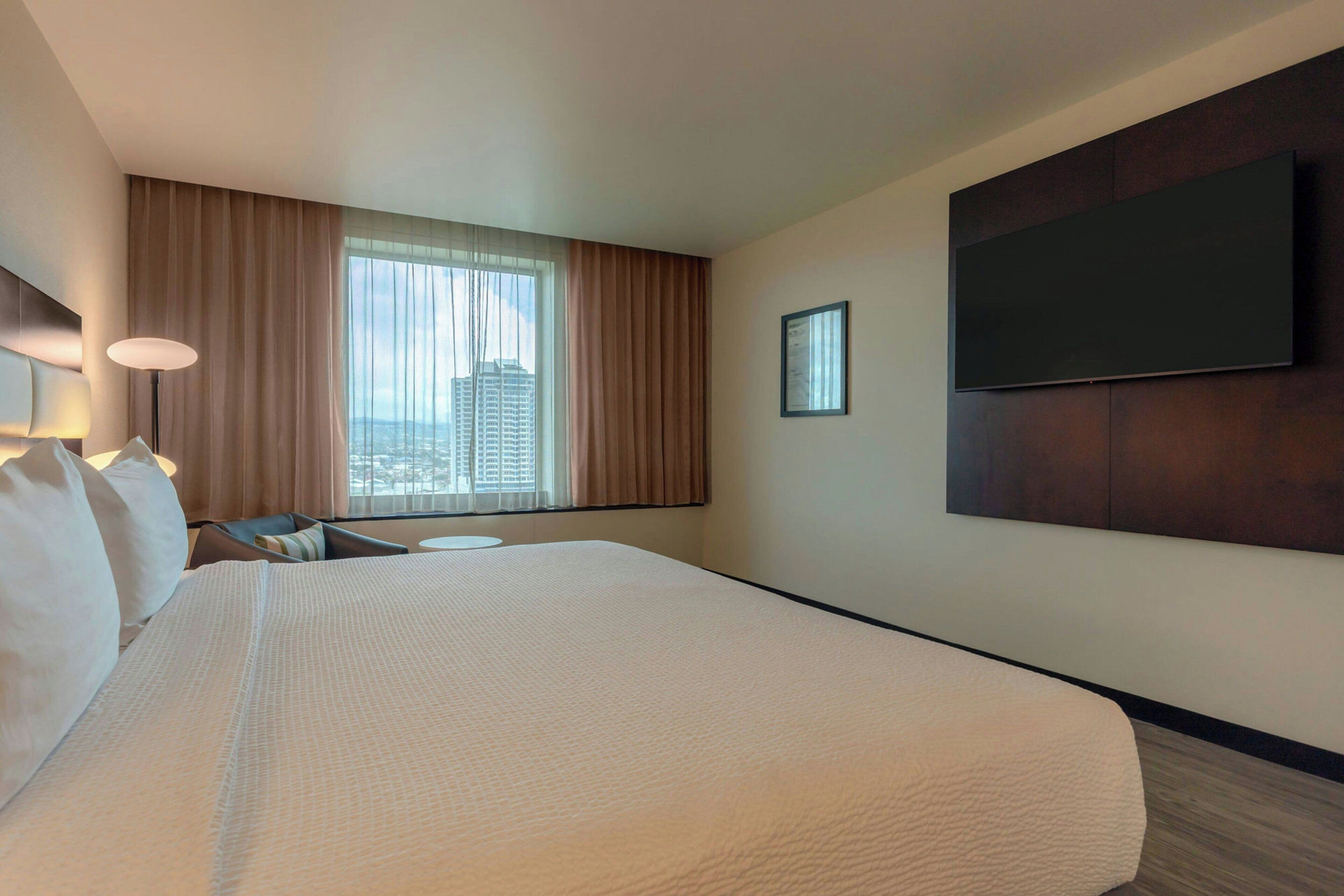 Habitación de hotel con una cama grande, un televisor de pantalla plana en la pared, una ventana con cortinas y vistas a la ciudad.