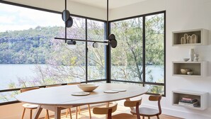 Zona pranzo con tavolo rotondo, sedie e vista su un lago e alberi attraverso ampie finestre.
