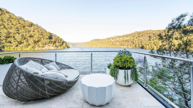 Un balcone con una sedia in vimini, un tavolo bianco e piante in vaso che si affaccia su un lago con colline sullo sfondo.