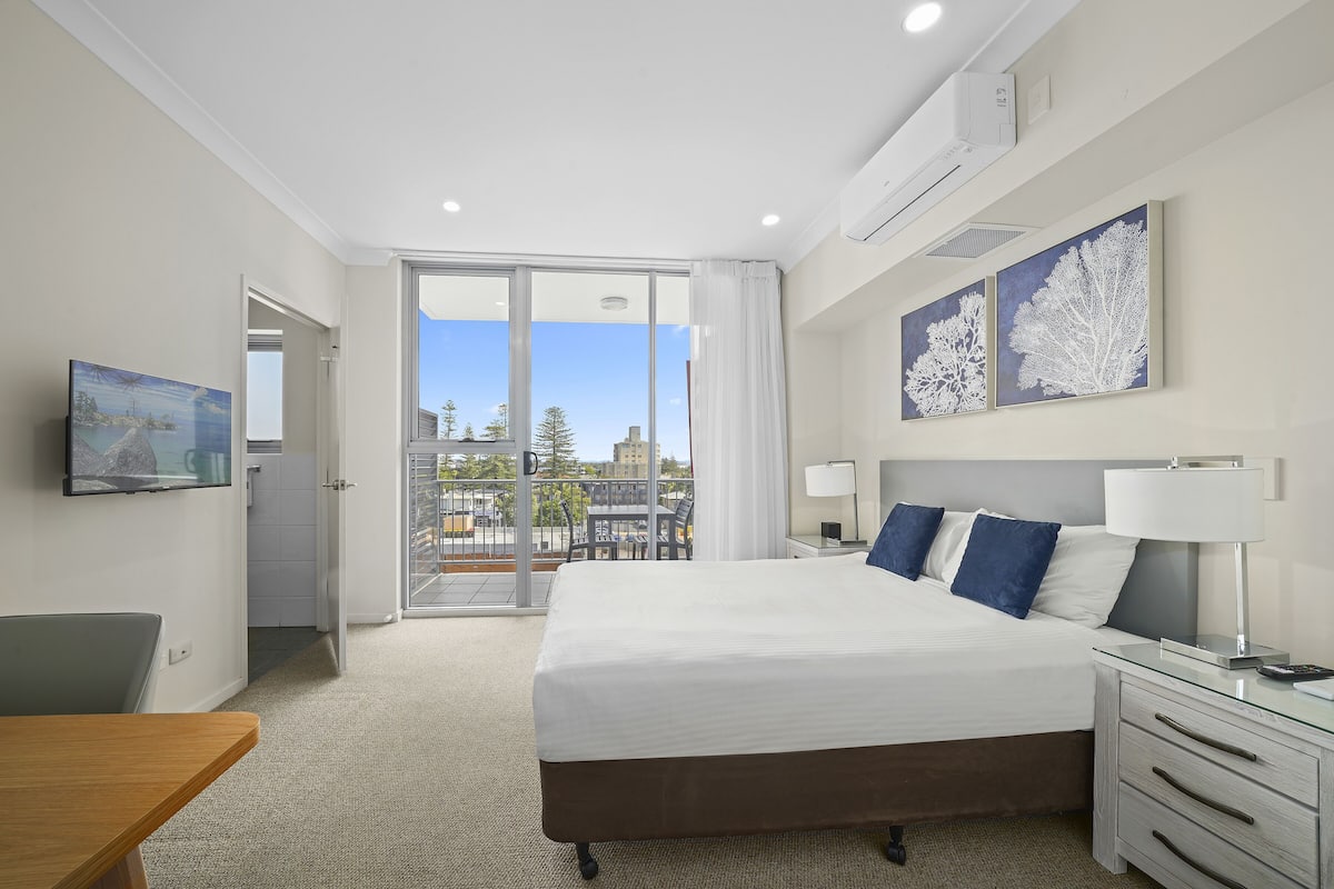 A hotel room with a large bed, a desk, a chair, a TV, and a balcony with a view of trees and buildings.