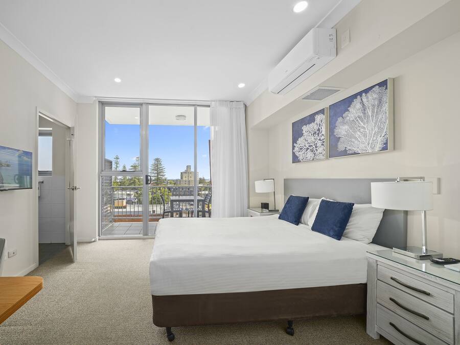 Een hotelkamer met een groot bed, een bureau, een stoel, een televisie en een balkon met uitzicht op bomen en gebouwen.