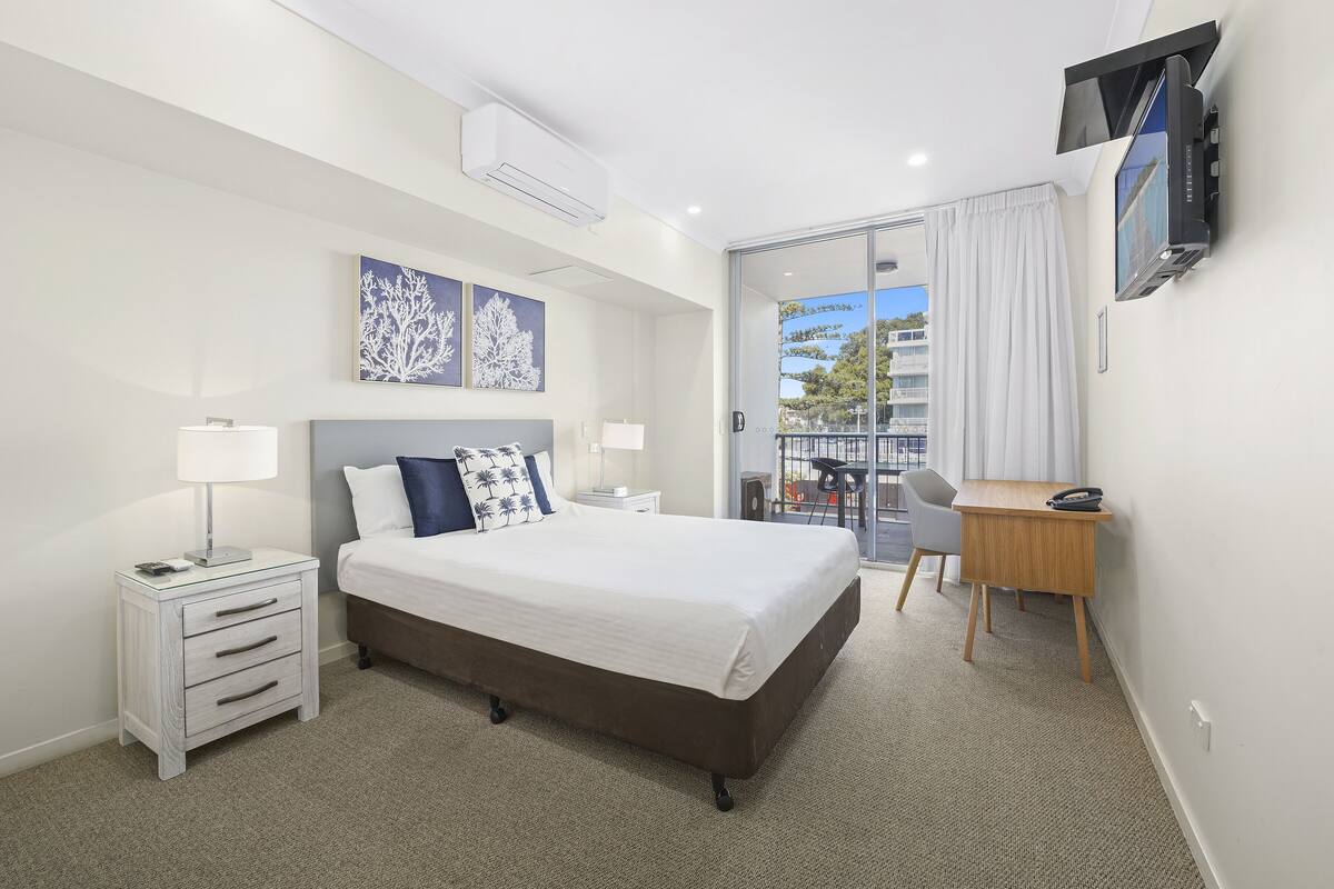 A hotel room with a bed, bedside tables, a desk, a chair, a TV, and a balcony with a view.