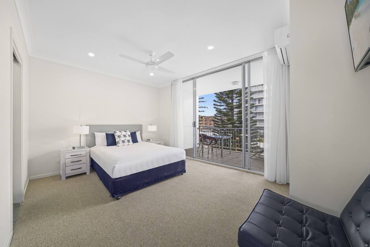 A hotel room with a large bed, a ceiling fan, a nightstand, a desk, and a view of the outside balcony.