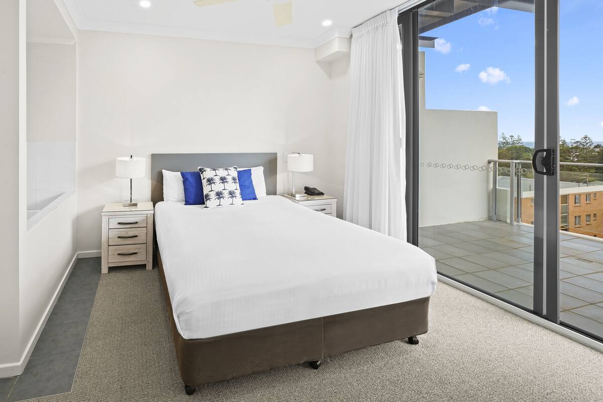 A modern hotel room with a large bed, bedside tables, a nightstand, and a view of the city through floor-to-ceiling windows.