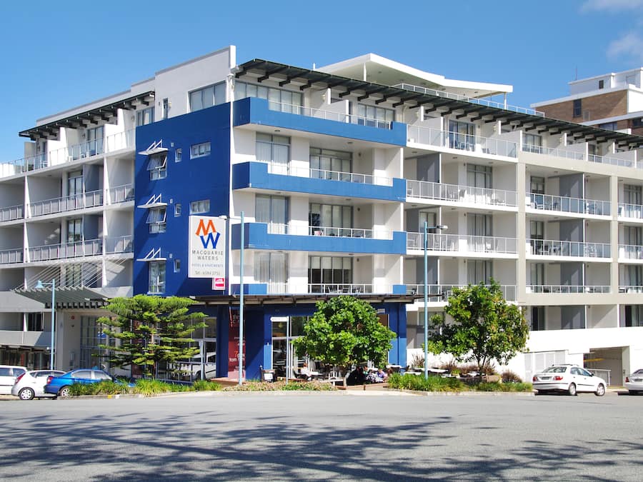 A multi-story hotel with balconies, a visible sign for 'MAQUARIE WATERS', and a parking area with cars.