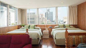 Habitación de hotel con dos camas, una silla roja, una mesita y ventanas amplias con vistas a la ciudad.