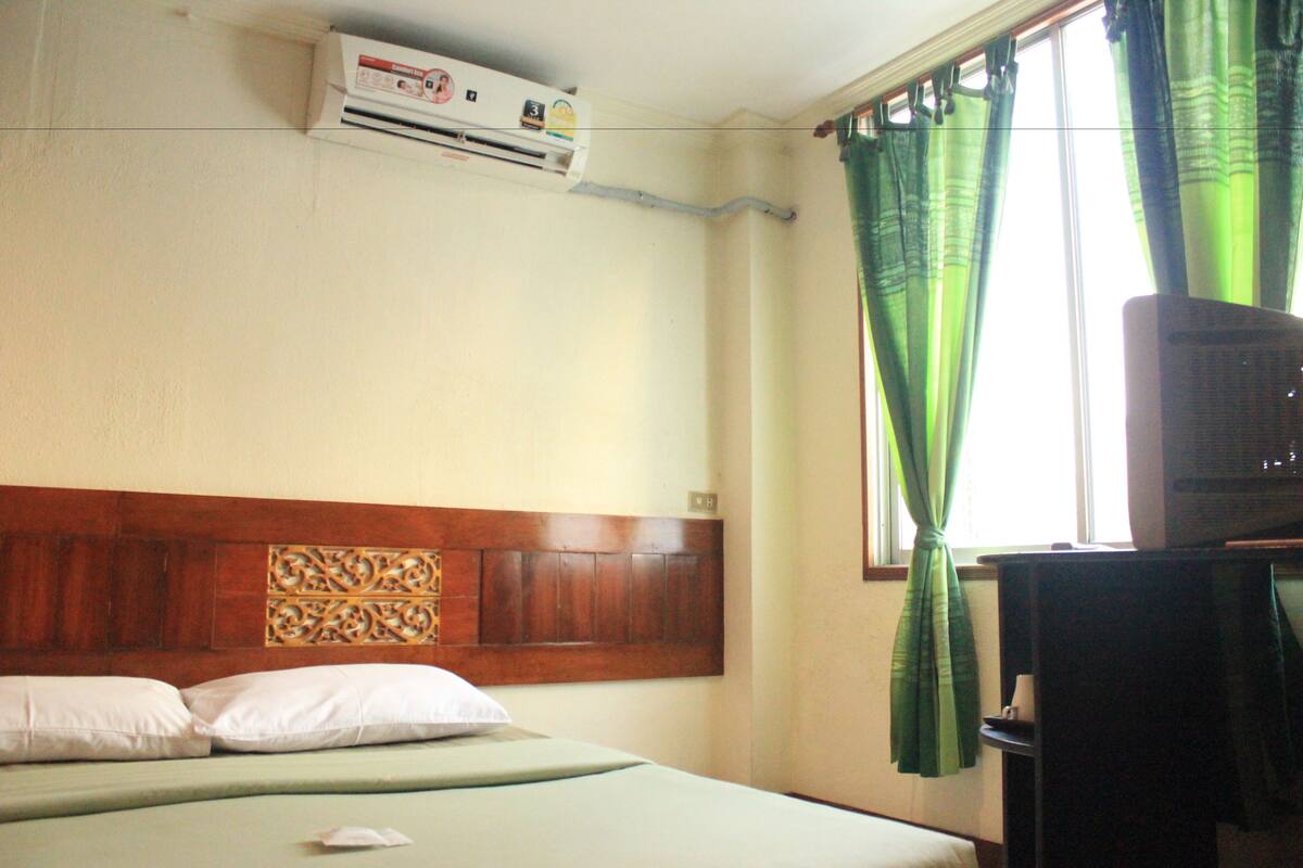 A hotel room with a bed, an air conditioning unit, a television, and green curtains.
