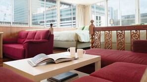 Una camera d'albergo moderna con un divano rosso, un tavolino in legno con una rivista e un libro, una tazza da caffè bianca e un letto con una coperta verde.