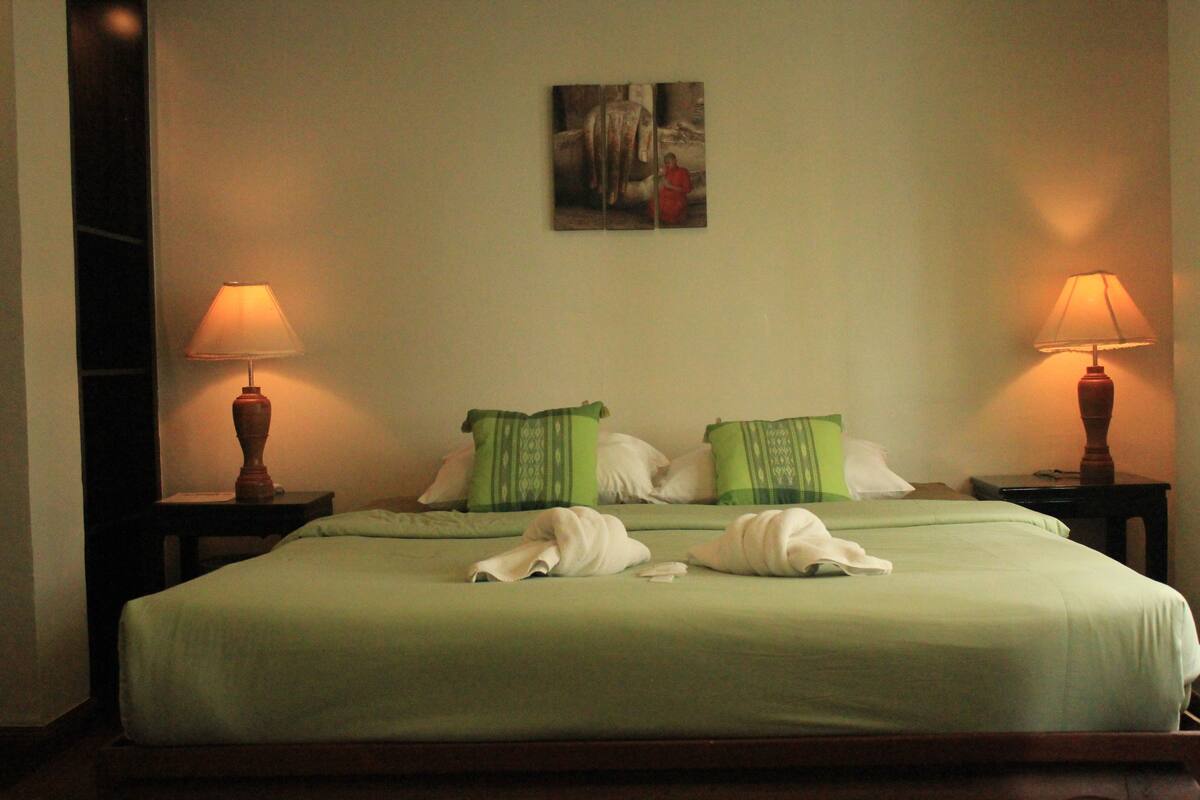 A neatly made bed with green and white bedding, two bedside lamps, and a painting on the wall.