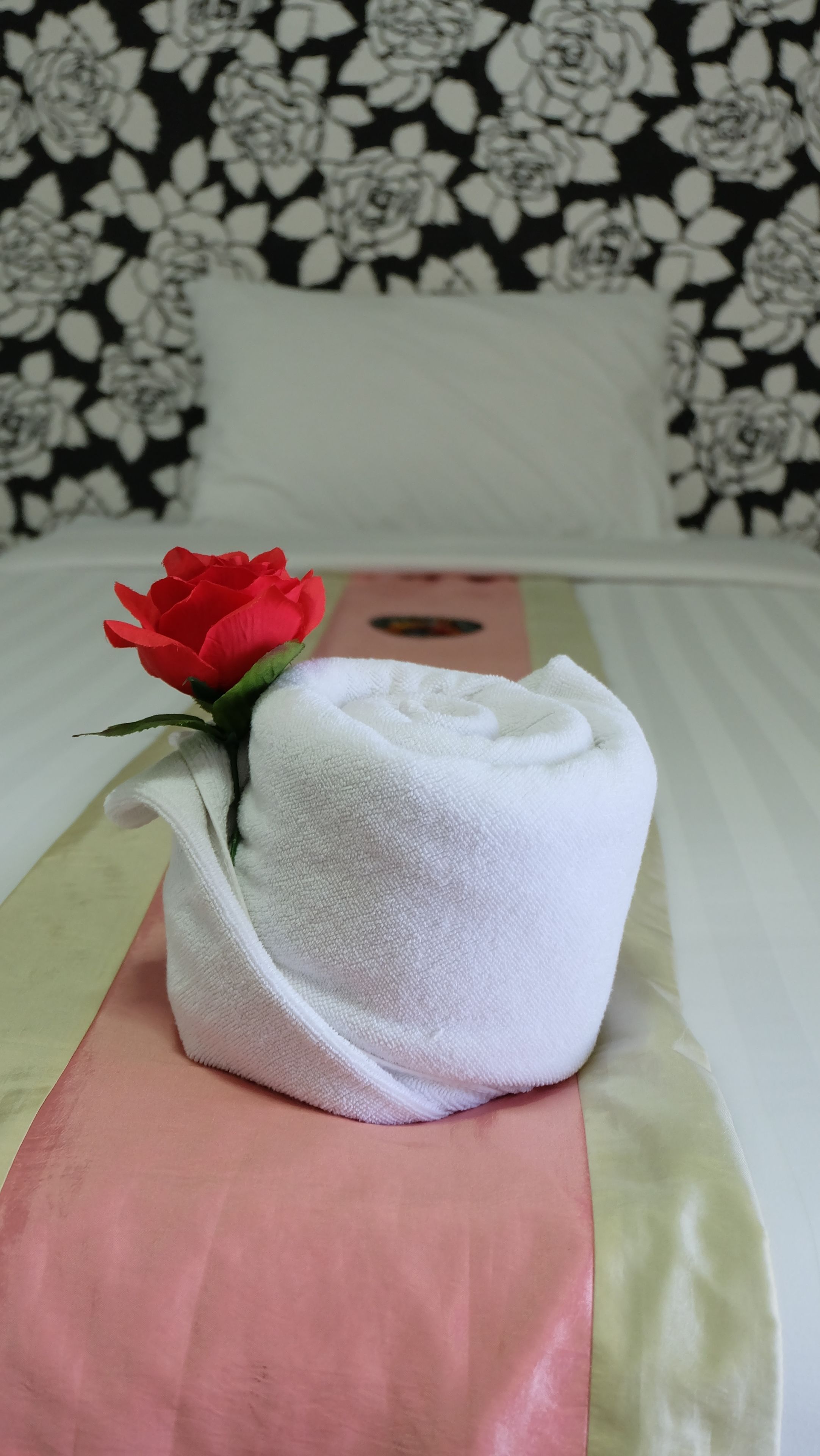 A neatly made bed with a white towel and a red rose placed on it.