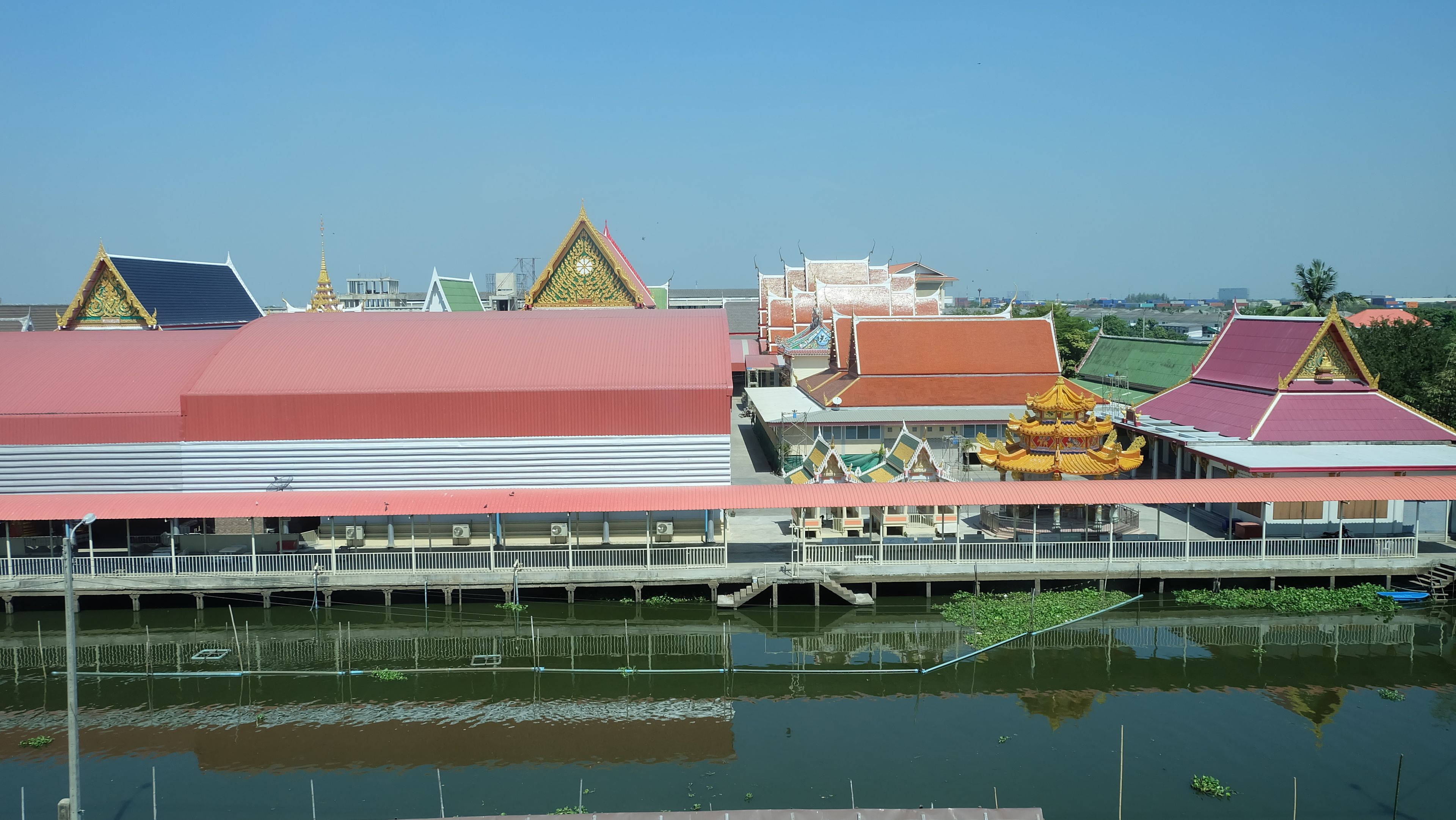 Un bâtiment traditionnel avec un toit rouge et des décorations dorées, un plan d’eau devant et un ciel dégagé.