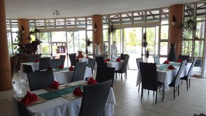 A restaurant with multiple tables, each set with white tablecloths, red napkins, and green placemats. The room has large windows allowing natural light and views of greenery outside. - Jet Over Hotel (Salelologa)