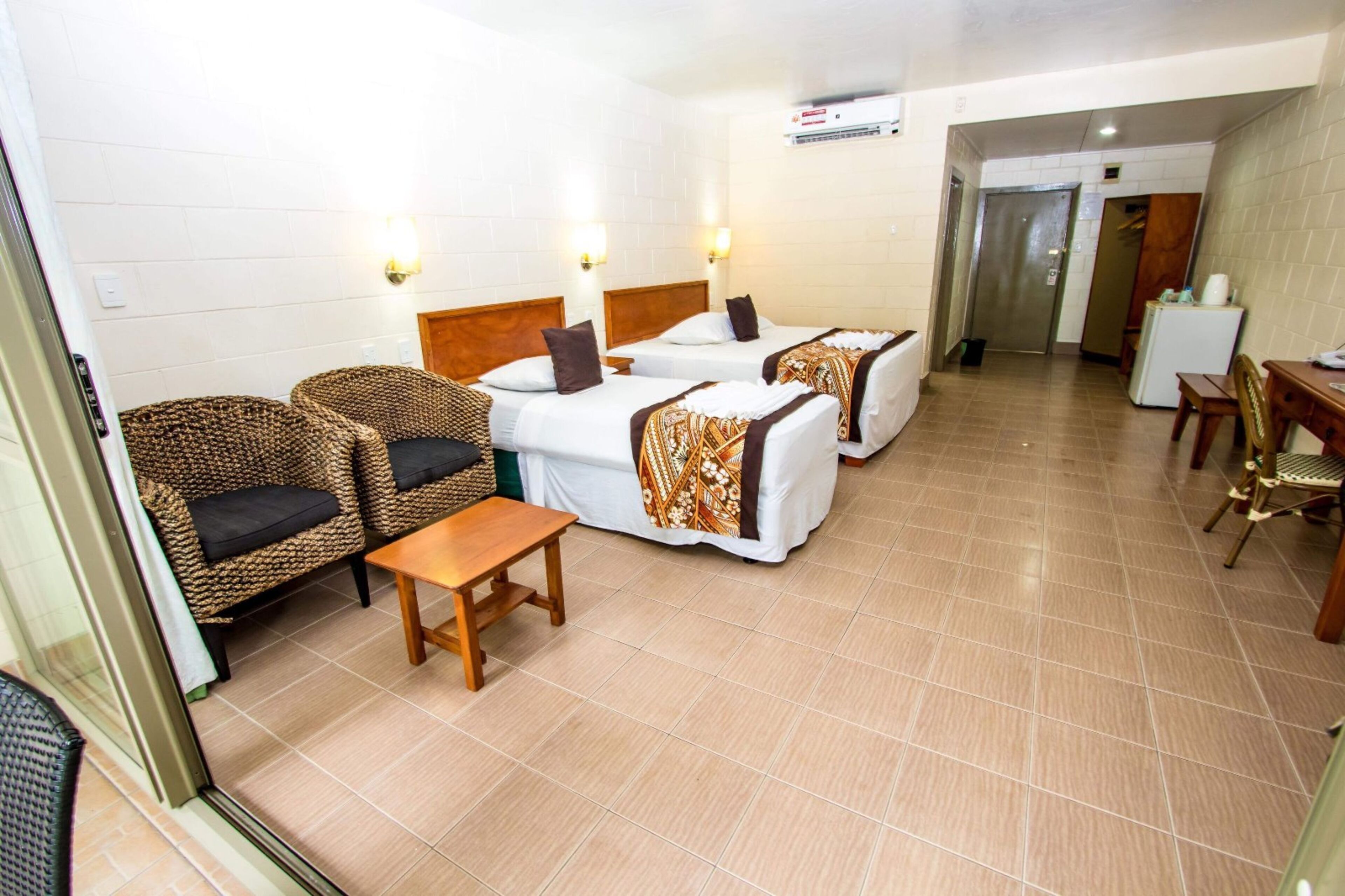 A hotel room with two beds, wicker chairs, a wooden table, and a small desk.