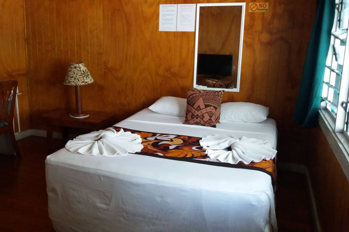 A hotel room with a wooden wall, a bed with a floral bedspread, a nightstand with a lamp, a chair, and a window with a view.