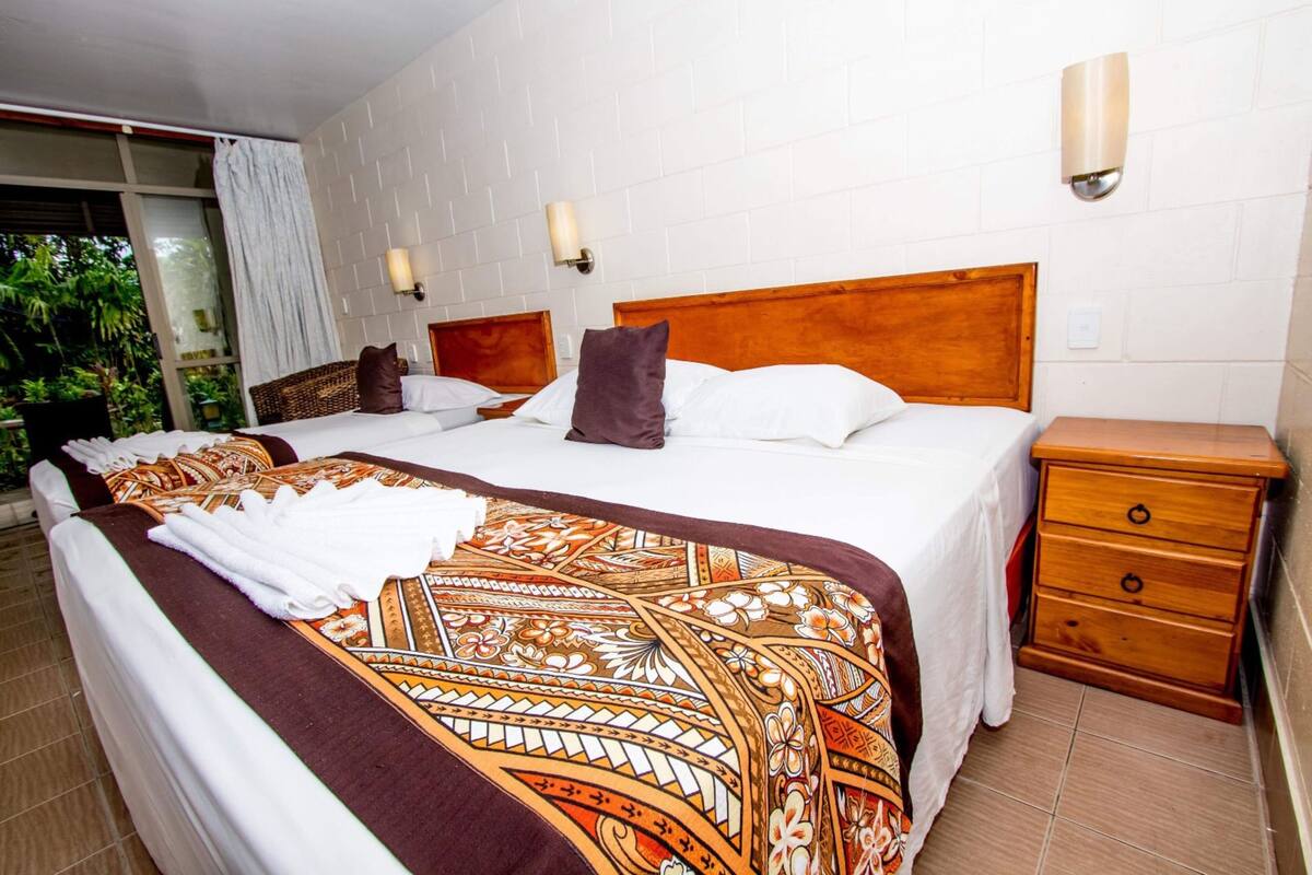 A hotel room with two beds, a wooden headboard, a nightstand with drawers, and a view of greenery outside.