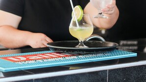 En bardisk med en avlång yta, två glas med en gulaktig drink garnerad med lime, och en person i bakgrunden som bär en svart t-shirt med text på ryggen.