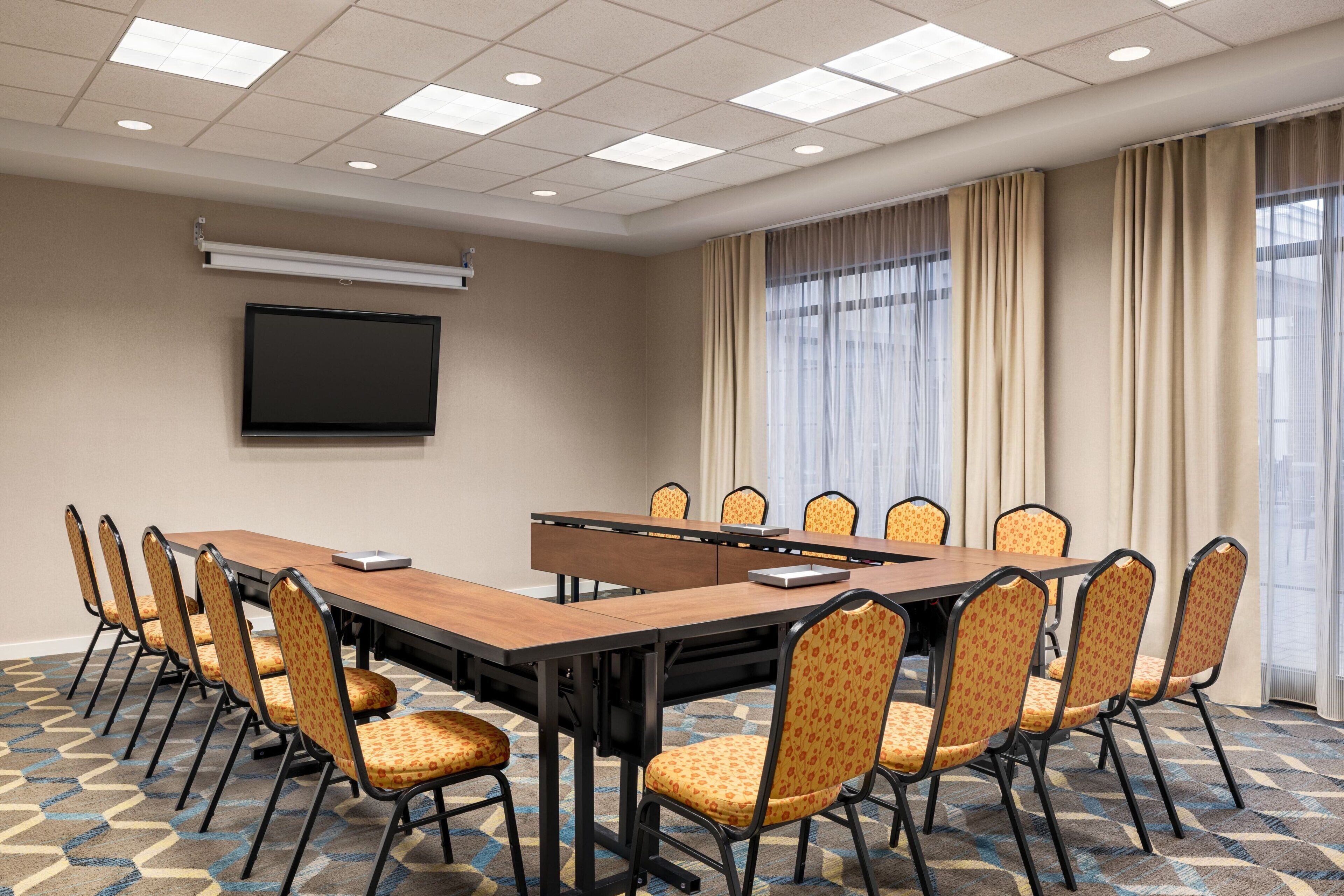 A conference room with a long table, chairs, a flat-screen TV, and large windows with curtains.