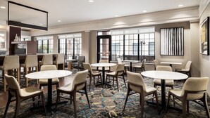 Un hall d’hôtel avec plusieurs tables et chaises rondes, de grandes fenêtres et un sol recouvert de moquette.