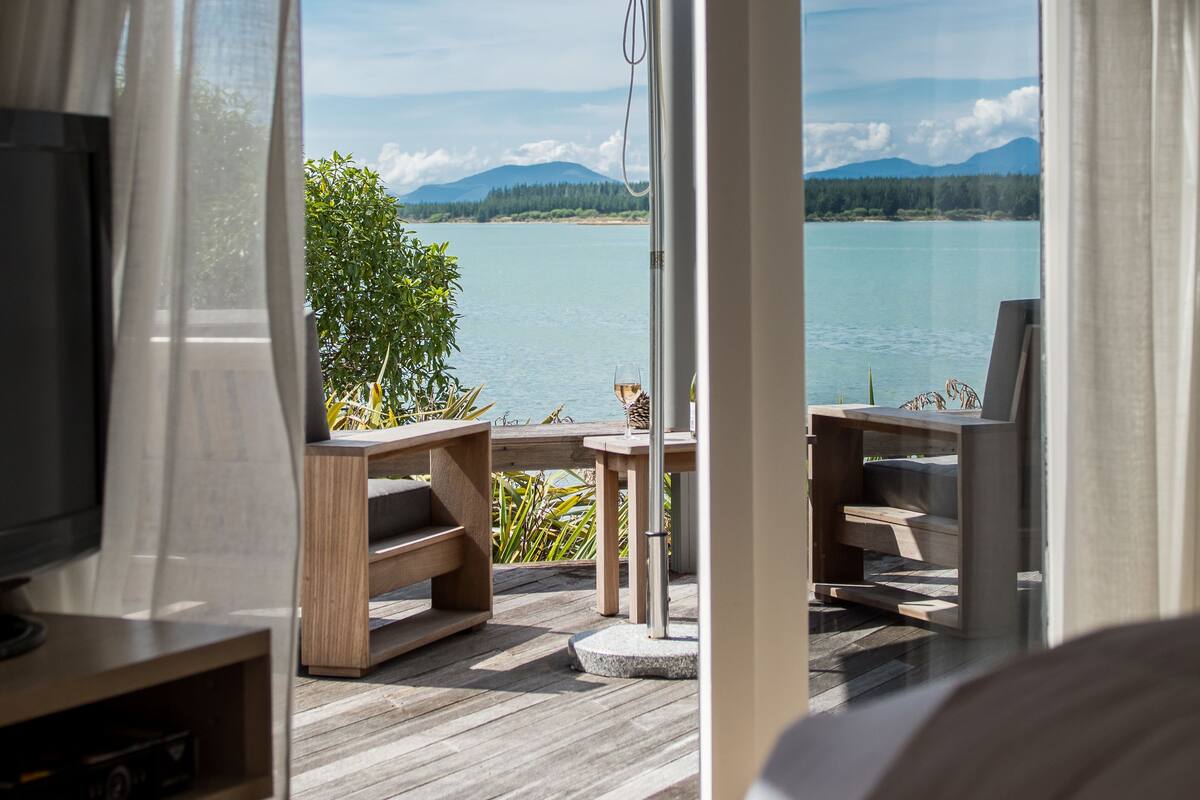 A room with a large window overlooking a lake, a wooden deck, and a table with a glass of wine.
