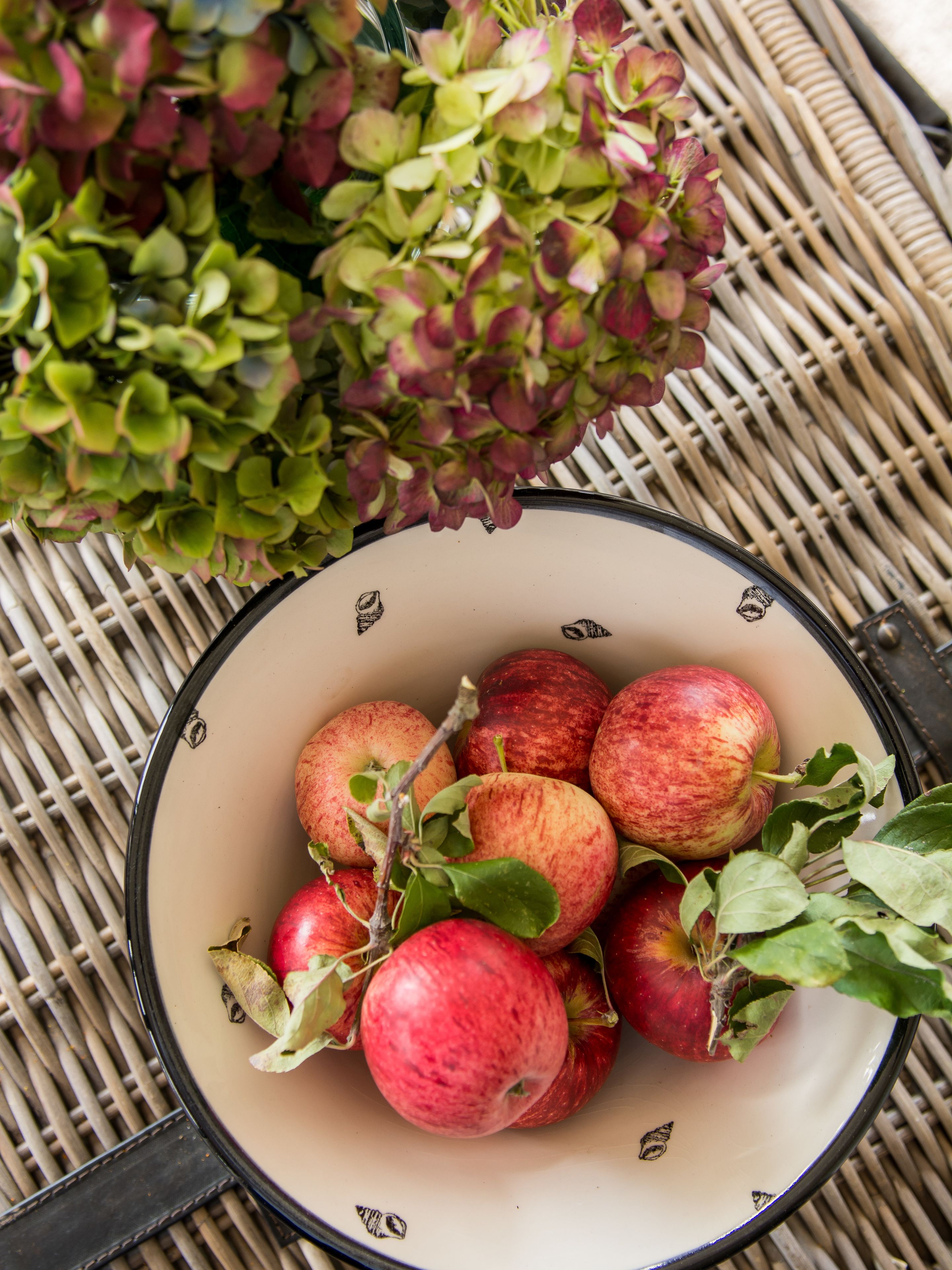 A bowl of apples and a bunch of hydrangeas on a woven surface.