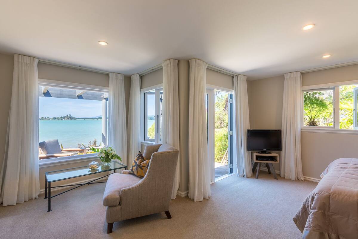 A hotel room with a large window offering a view of the sea, a comfortable armchair, a television, and a bed.