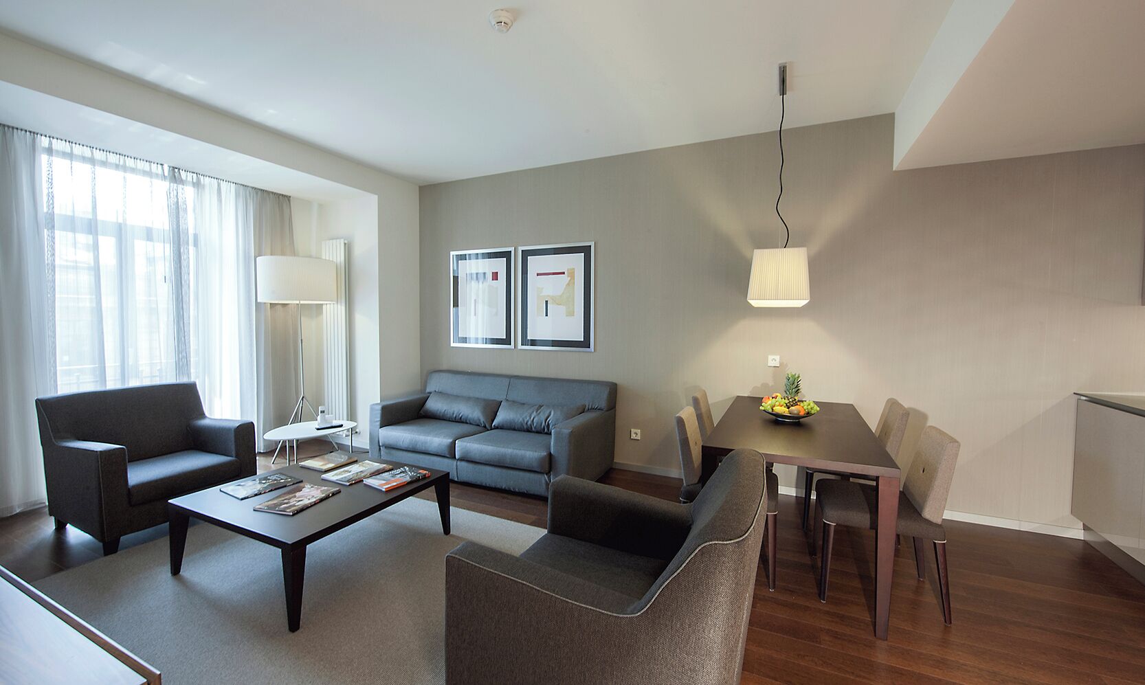 A modern living room with a sofa, armchairs, a coffee table, and a dining table with chairs. There is a lamp, framed pictures on the wall, and a fruit bowl on the table.