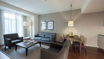 A modern living room with a sofa, armchairs, a coffee table, and a dining table with chairs. There is a lamp, framed pictures on the wall, and a fruit bowl on the table.