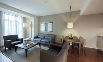 A modern living room with a sofa, armchairs, a coffee table, and a dining table with chairs. There is a lamp, framed pictures on the wall, and a fruit bowl on the table.