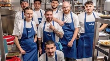Un grupo de personas en una cocina con electrodomésticos de acero inoxidable y una exhibición de alimentos.