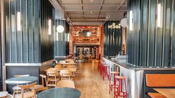 Un intérieur de restaurant moderne avec des tables en bois et des tabourets de bar rouges, un bar central et une vue sur la cuisine.