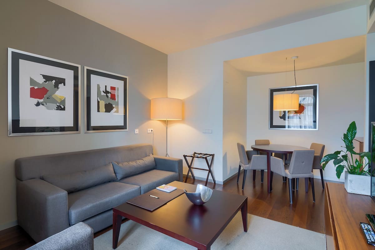 A modern living room with a grey sofa, a wooden coffee table, and a dining area with a table and chairs. There are two framed abstract artworks on the wall, a floor lamp, and a potted plant.