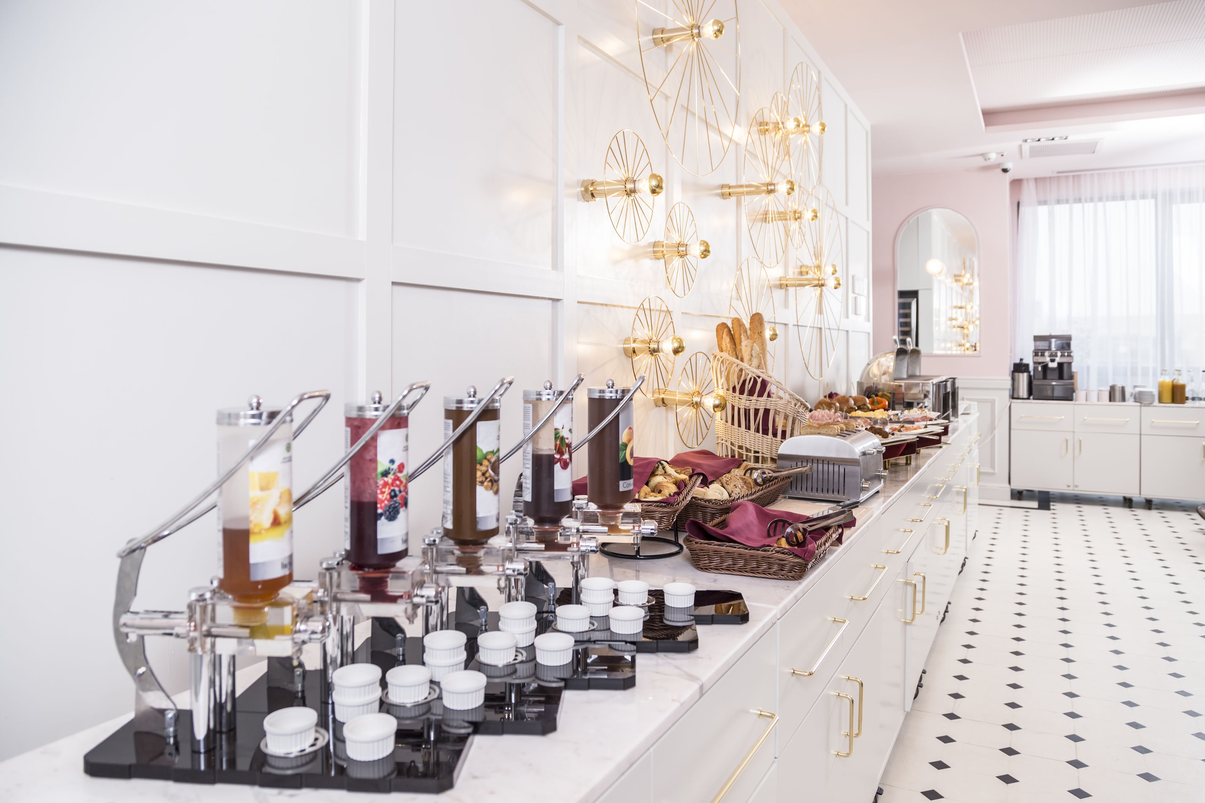 A breakfast buffet with various food items and beverages, a coffee machine, and a decorative wall with hanging lights.