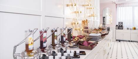 Un bufé de desayuno con diversas opciones de comida y bebidas, una máquina de café y una pared decorativa con luces colgantes.
