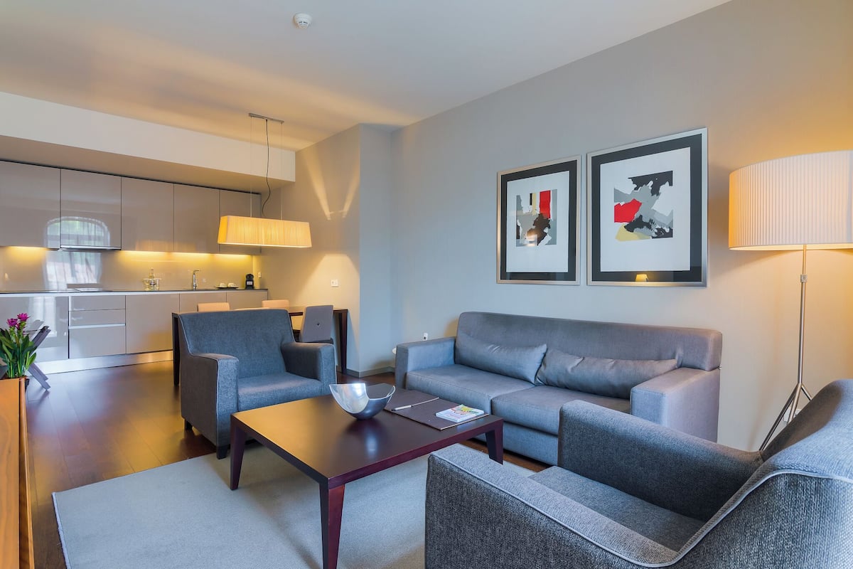 A modern living room with a grey sofa, armchairs, and a coffee table. There is a kitchen area with white cabinets and a lamp. Two framed artworks are on the wall.