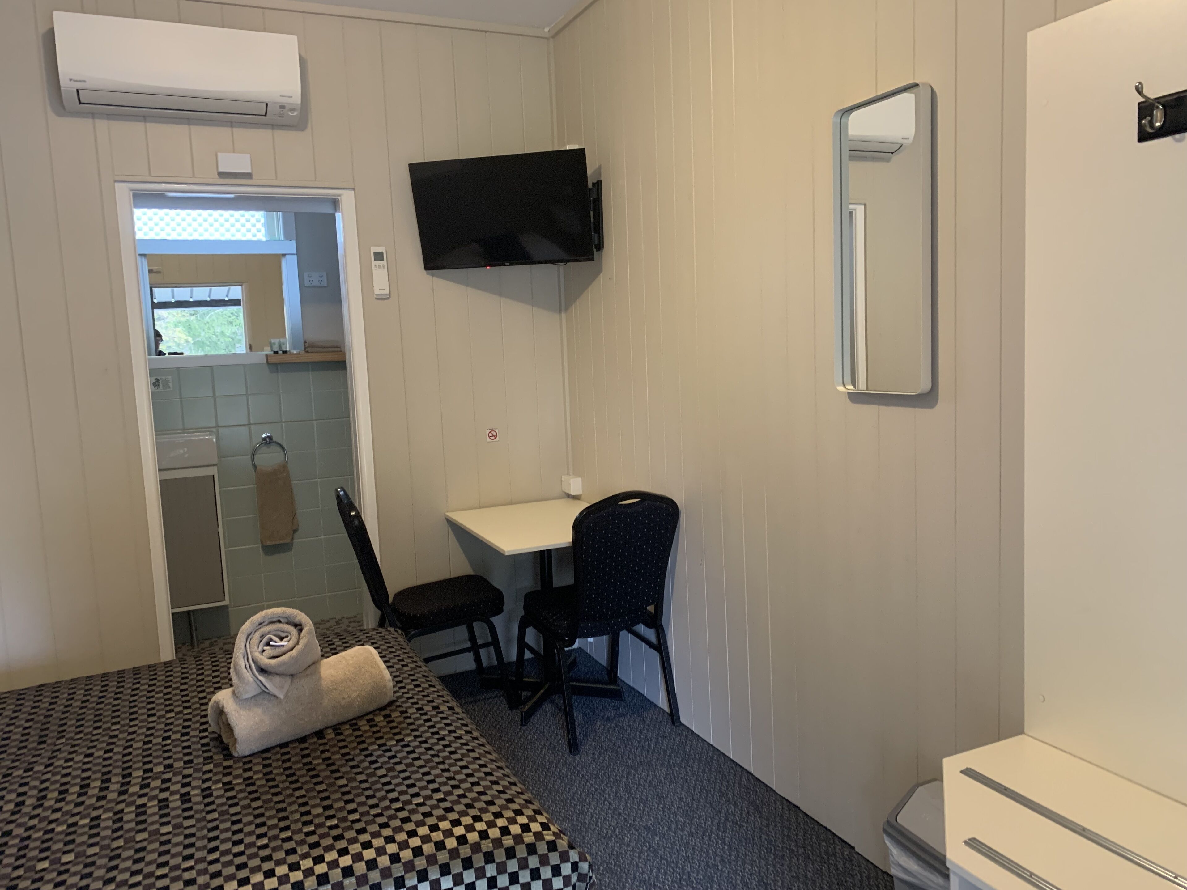 Una habitación pequeña y limpia con cama, una silla, un escritorio, un televisor y un espejo.