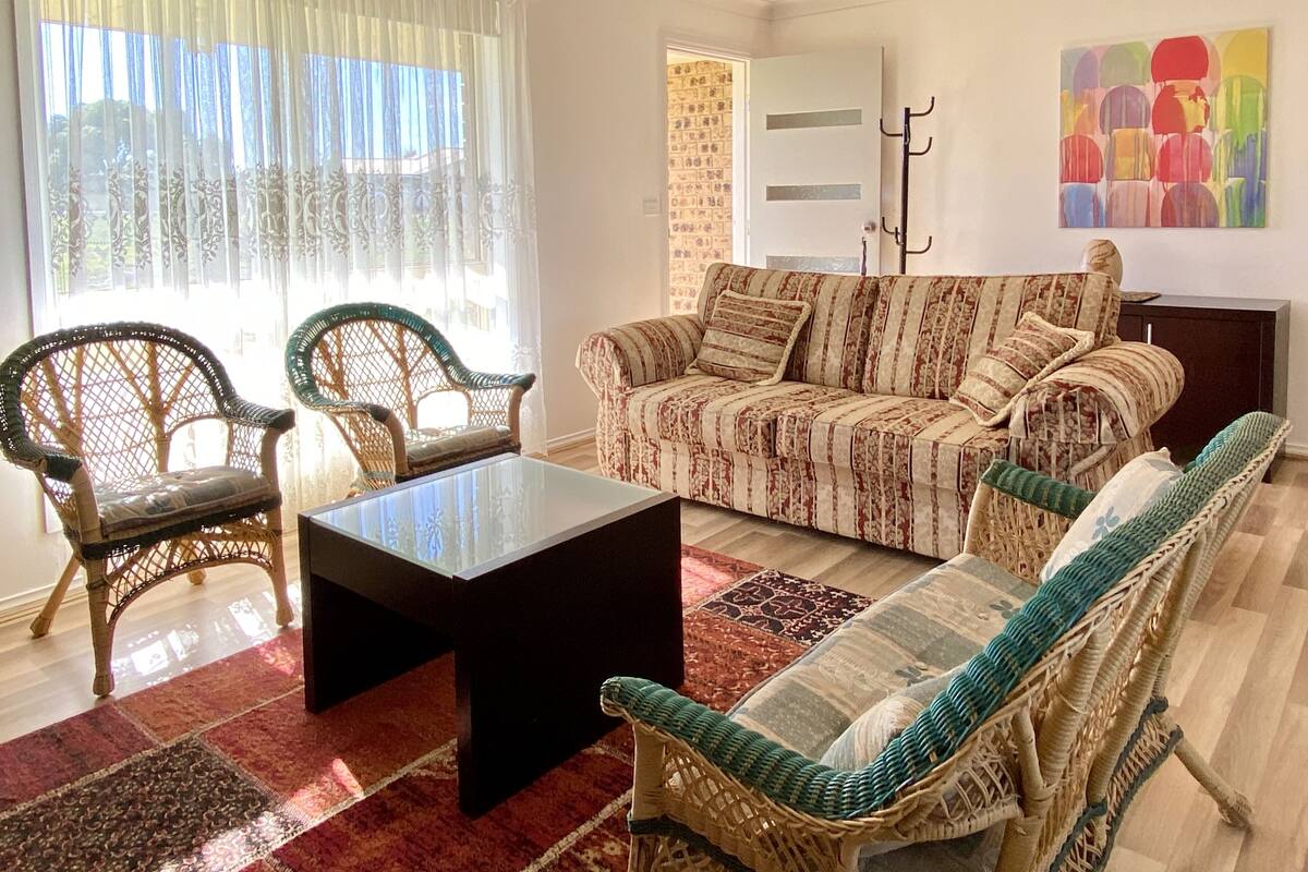 Una sala de estar con muebles de mimbre, un sofá estampado, una mesa de centro y un cuadro abstracto y colorido en la pared.