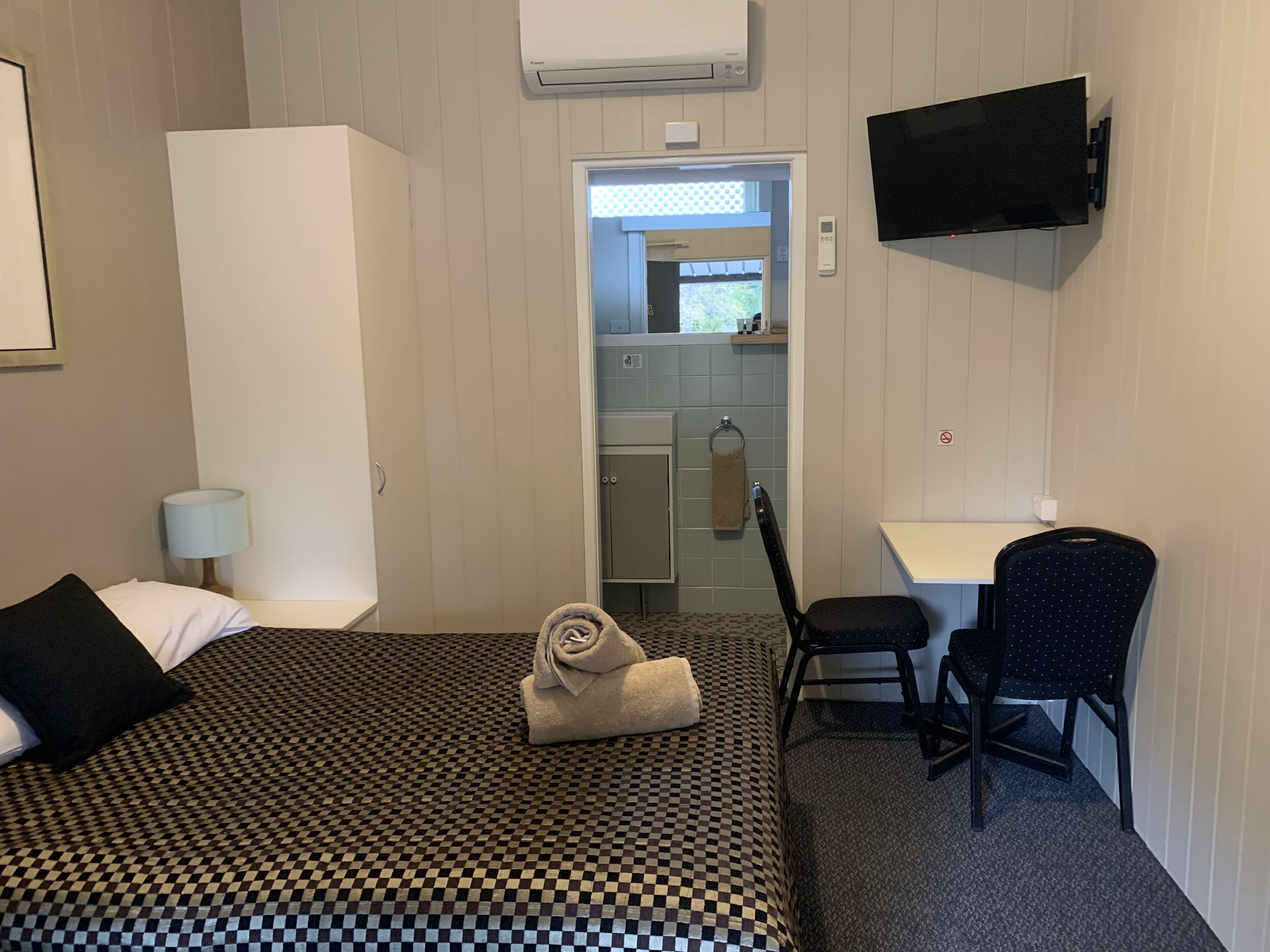 Una habitación de hotel con una colcha a cuadros, un escritorio con una silla, un televisor montado en la pared y un baño visible a través de una puerta abierta.